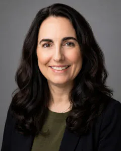 Headshot of a smiling woman with long dark wavy hair, wearing a dark blazer over a green top, against a neutral gray background.
