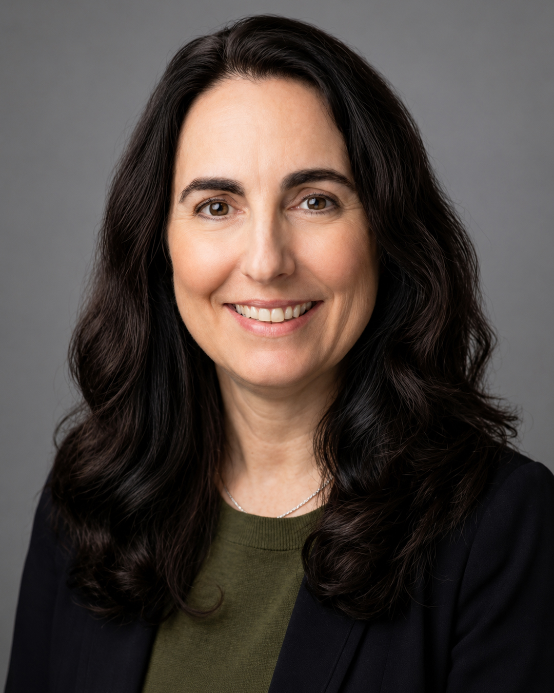 Headshot of a smiling woman with long dark wavy hair, wearing a dark blazer over a green top, against a neutral gray background.