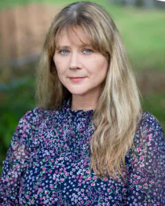 Headshot of a woman with long blonde hair and blue eyes, wearing a dark floral blouse, outdoors with a softly blurred green background.