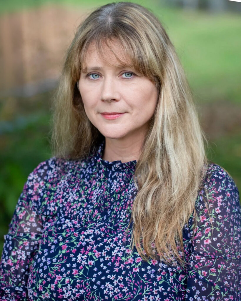 Headshot of a woman with long blonde hair and blue eyes, wearing a dark floral blouse, outdoors with a softly blurred green background.
