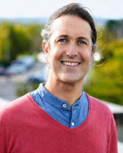 Smiling person with long hair pulled back, wearing a coral-red sweater over a blue button-up shirt, standing outdoors against a softly blurred background of trees and buildings. The portrait is brightly lit with natural light and framed as a professional headshot, with the subject looking directly at the camera.