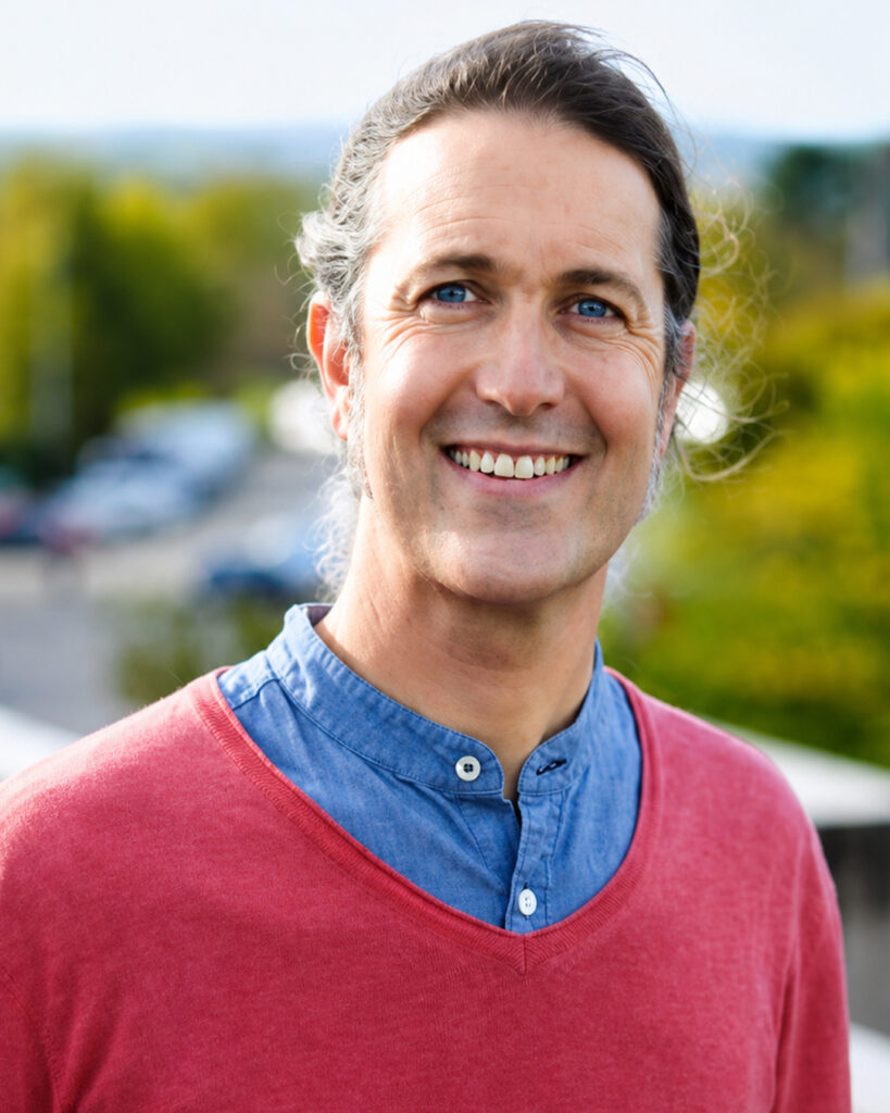Smiling person with long hair pulled back, wearing a coral-red sweater over a blue button-up shirt, standing outdoors against a softly blurred background of trees and buildings. The portrait is brightly lit with natural light and framed as a professional headshot, with the subject looking directly at the camera.