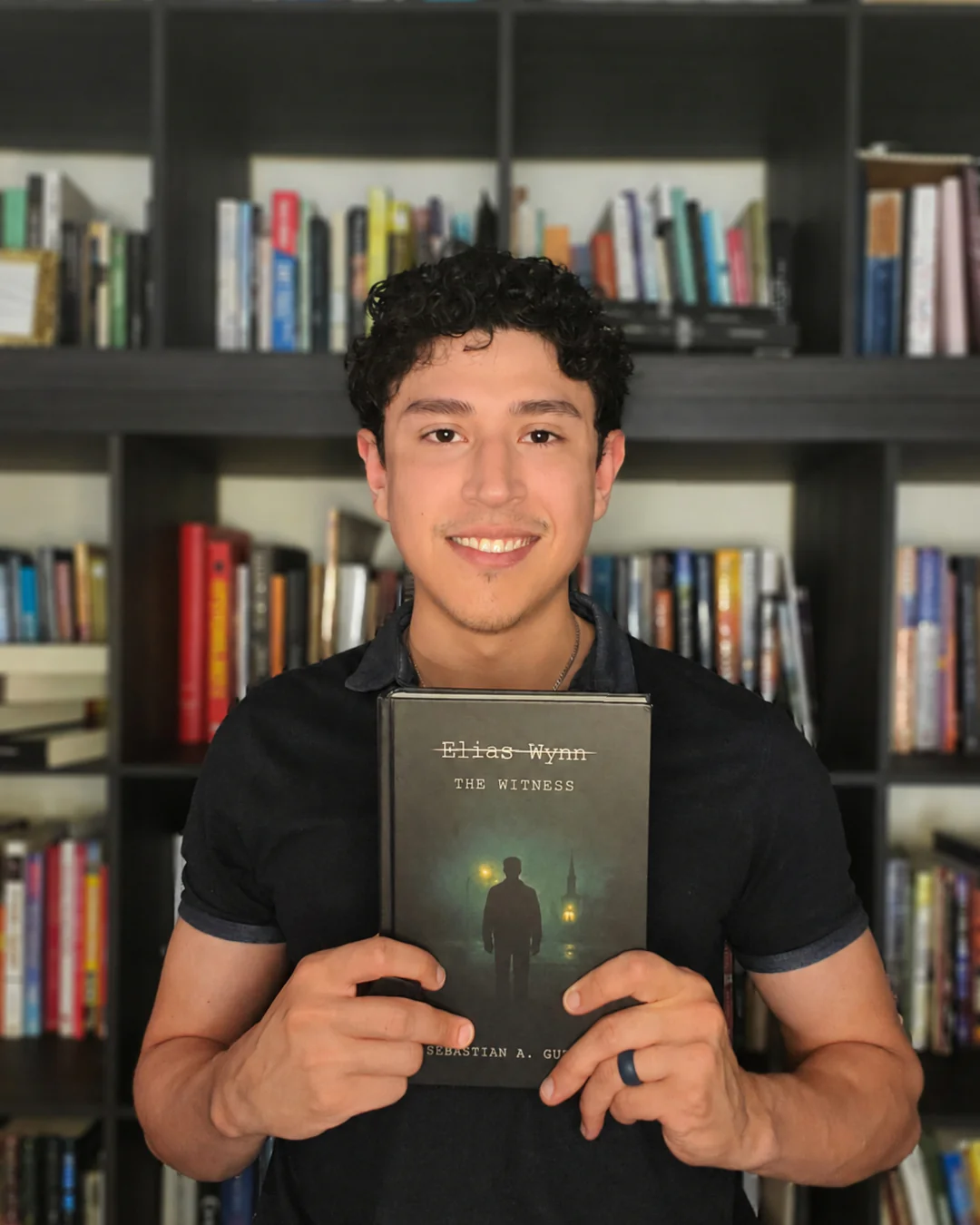 Sebastian pictured in front of book case holding "Elias Wynn THE WITNESS," a book he wrote.