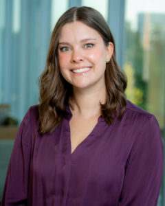 Professional headshot of a smiling woman with shoulder-length wavy brown hair wearing a purple blouse and stud earrings, posed in front of a softly blurred modern office background with large windows.
