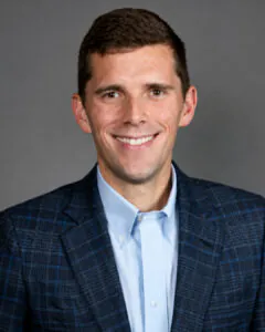 Professional headshot of a smiling man with short dark brown hair, wearing a light blue dress shirt and a navy blue plaid blazer. He is facing the camera against a neutral gray studio background with even lighting and a friendly, approachable expression.