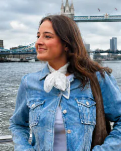 **Alt text:** A smiling young woman with long brown hair stands on a boat along the River Thames in London, wearing a light blue denim jacket over a white top with a sheer white neck scarf. She looks to the side with Tower Bridge prominently visible behind her under an overcast sky.