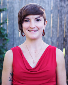 Portrait of a smiling woman with short brown hair, wearing a sleeveless red top, matching red hoop earrings, and a small pendant necklace. She is standing outdoors in front of a wooden fence and trees, with soft natural light and a blurred green background.
