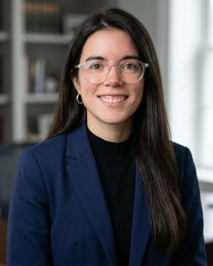 Professional headshot of a smiling woman with long dark brown hair and clear-framed glasses, wearing a navy blazer over a black top. She is facing the camera in a bright office setting with softly blurred bookshelves and windows in the background.