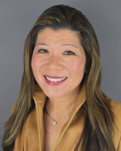 Professional headshot of a smiling woman with long highlighted brown hair, wearing a tan collared jacket and a delicate pendant necklace. She is facing the camera against a neutral gray studio background with a warm, approachable expression.