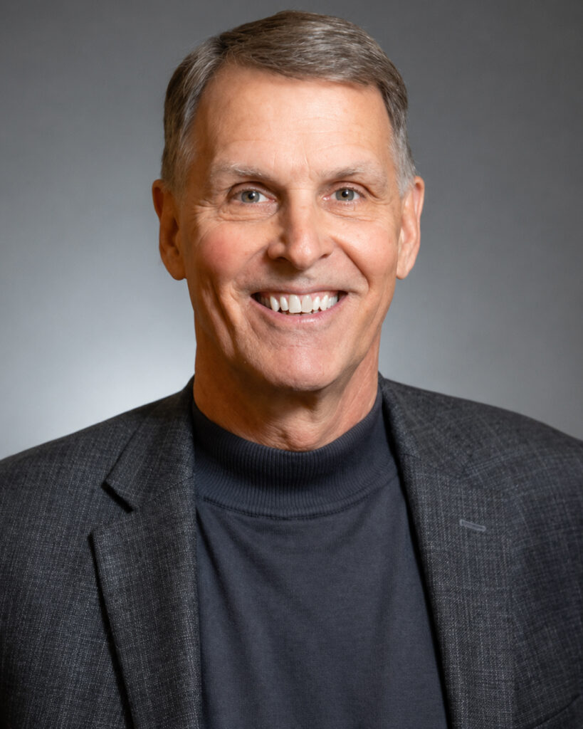 Head-and-shoulders professional portrait of a smiling man with short gray hair wearing a dark gray blazer over a charcoal mock-neck shirt. He faces the camera against a softly lit gray studio background.