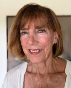 Professional headshot of a smiling woman with short light brown hair and bangs wearing a white top, photographed indoors against a softly blurred neutral background.