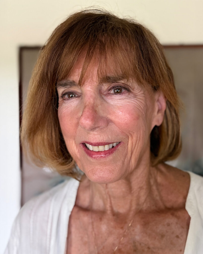 Professional headshot of a smiling woman with short light brown hair and bangs wearing a white top, photographed indoors against a softly blurred neutral background.