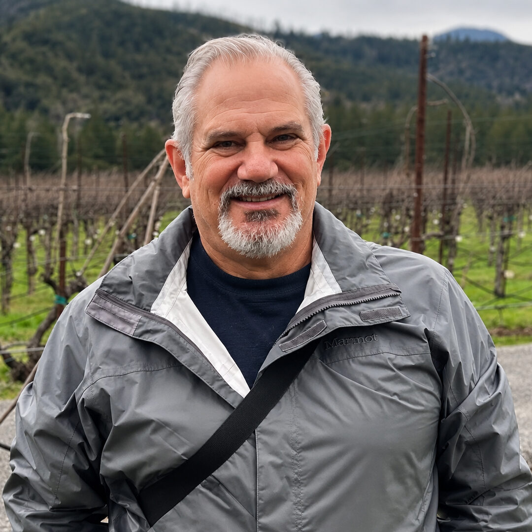 Man with gray hair and a goatee smiling outdoors in a vineyard, with tree-covered mountains in the background.