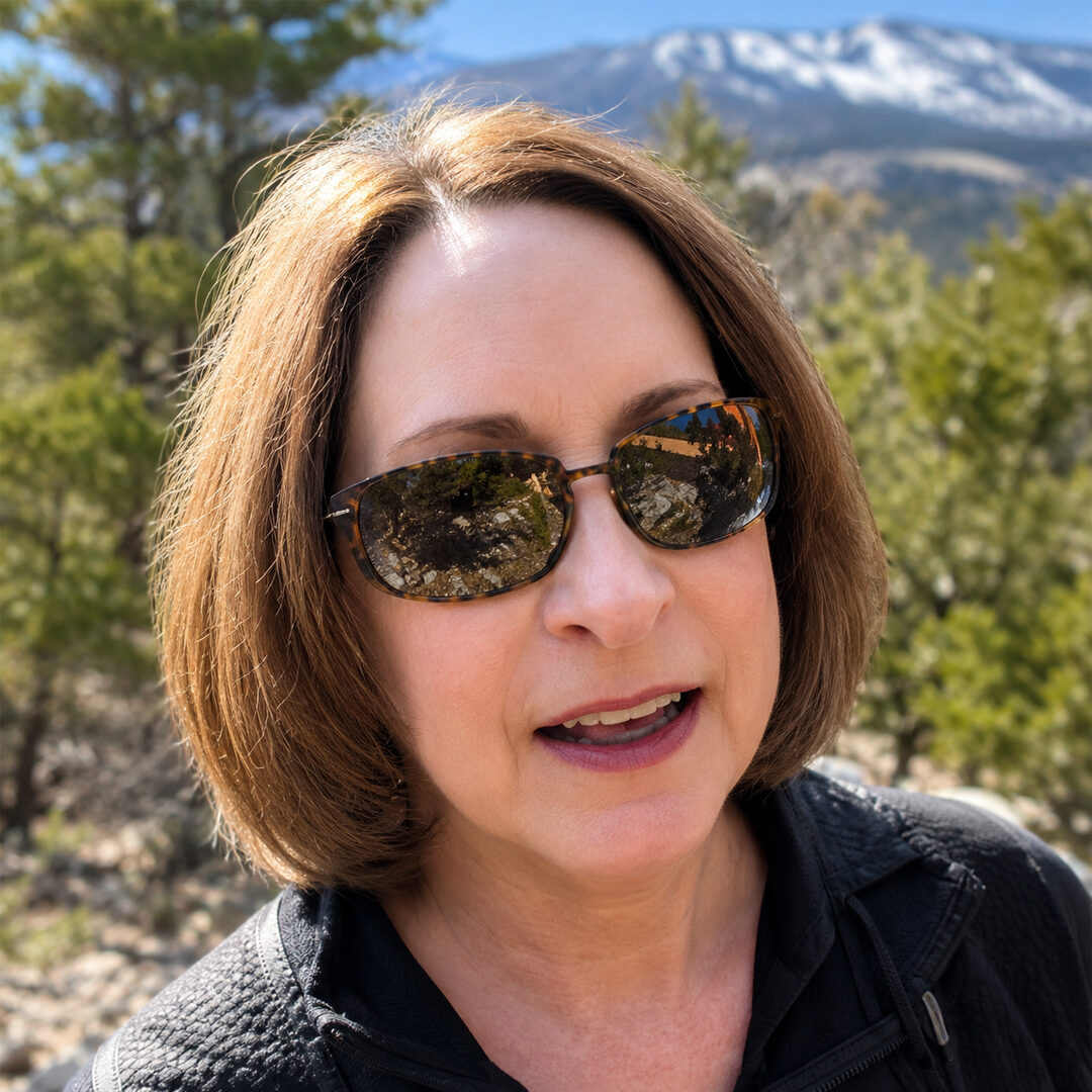 Woman with a brown bob haircut and sunglasses smiling outdoors, with trees and a snow-capped mountain in the background.