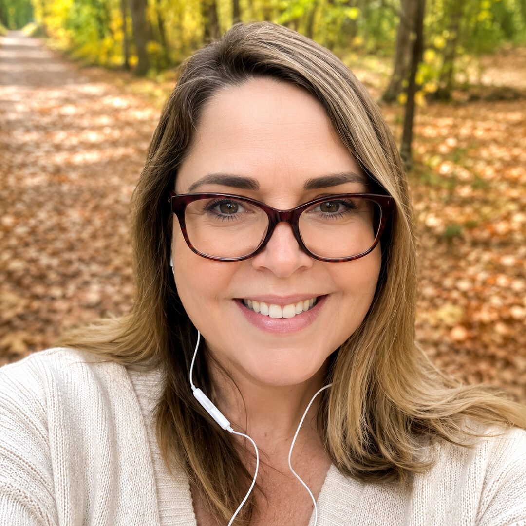 Smiling woman with long brown hair and glasses wearing a cream sweater and earbuds on a wooded trail covered with fallen leaves.