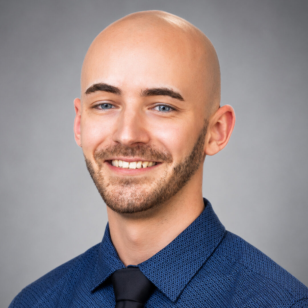 Smiling bald man with a short beard wearing a blue patterned dress shirt and dark tie against a gray studio background.