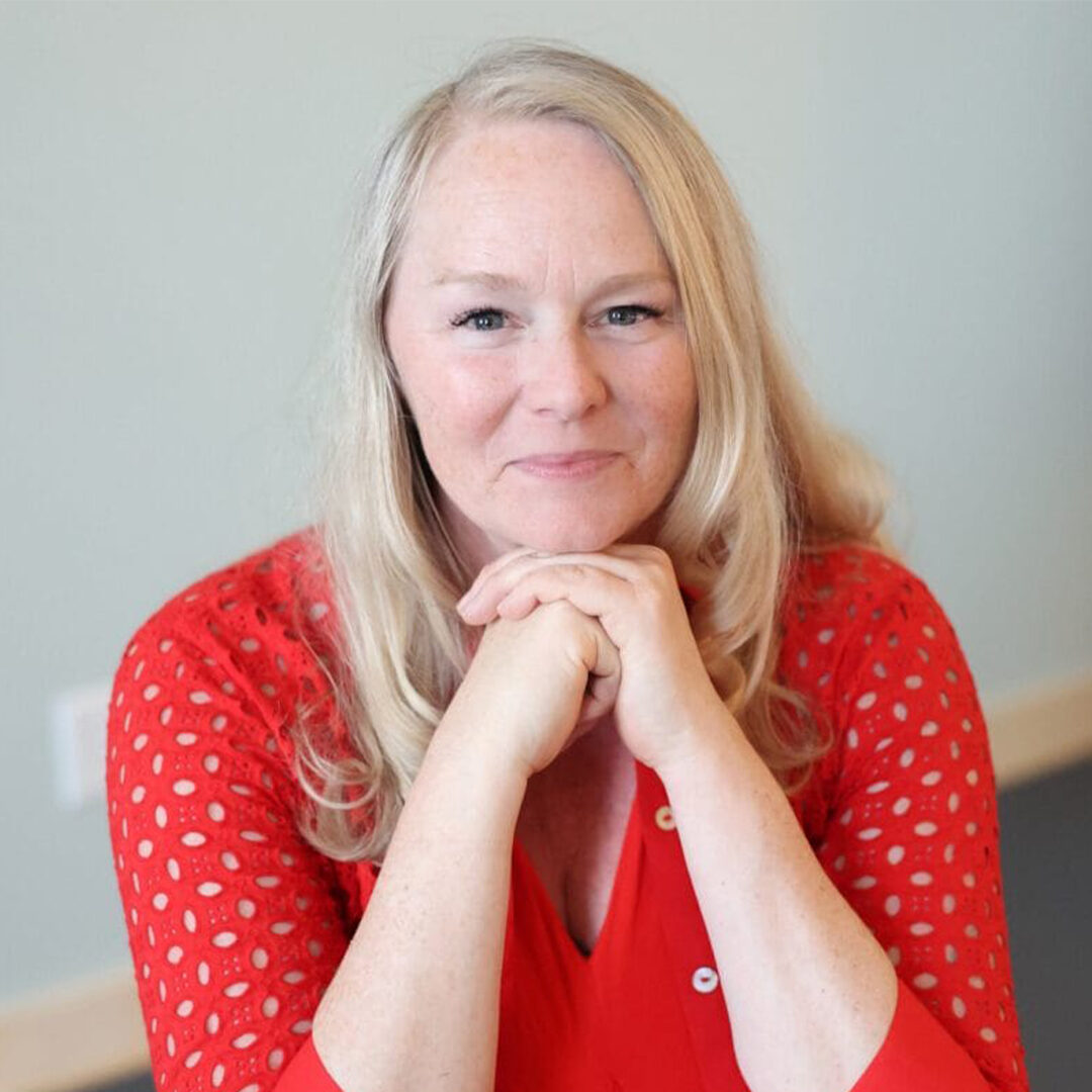 Blonde woman in a bright red blouse seated with her hands resting beneath her chin against a neutral background.