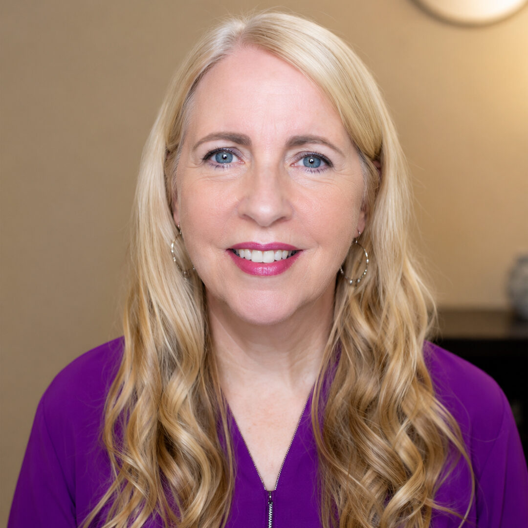 Smiling blonde woman wearing a purple blouse, photographed against a softly blurred indoor background.