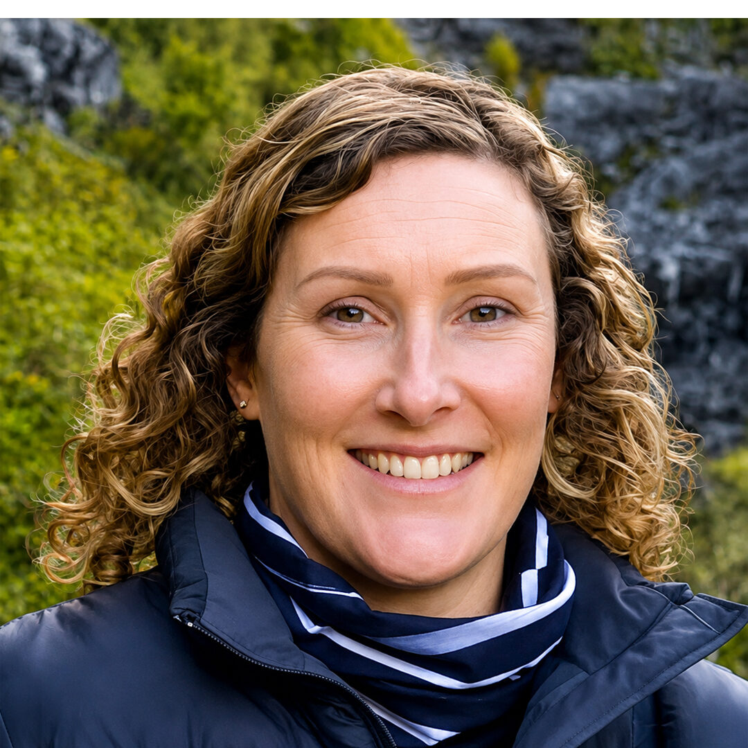 Smiling woman with curly brown hair wearing a navy jacket and striped scarf outdoors in a rocky, wooded setting.