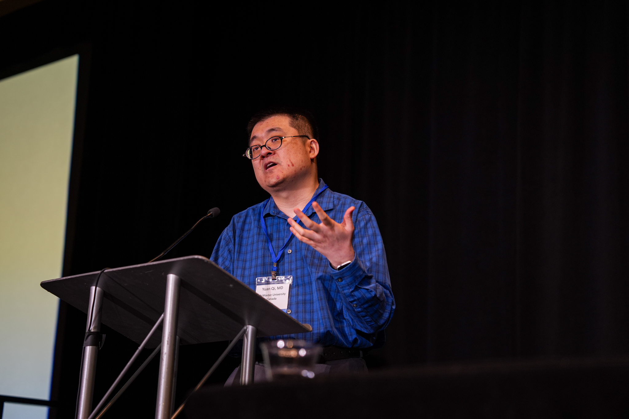 A physician speaks at a podium during the International Vasculitis Symposium, gesturing as he addresses the audience from the stage.