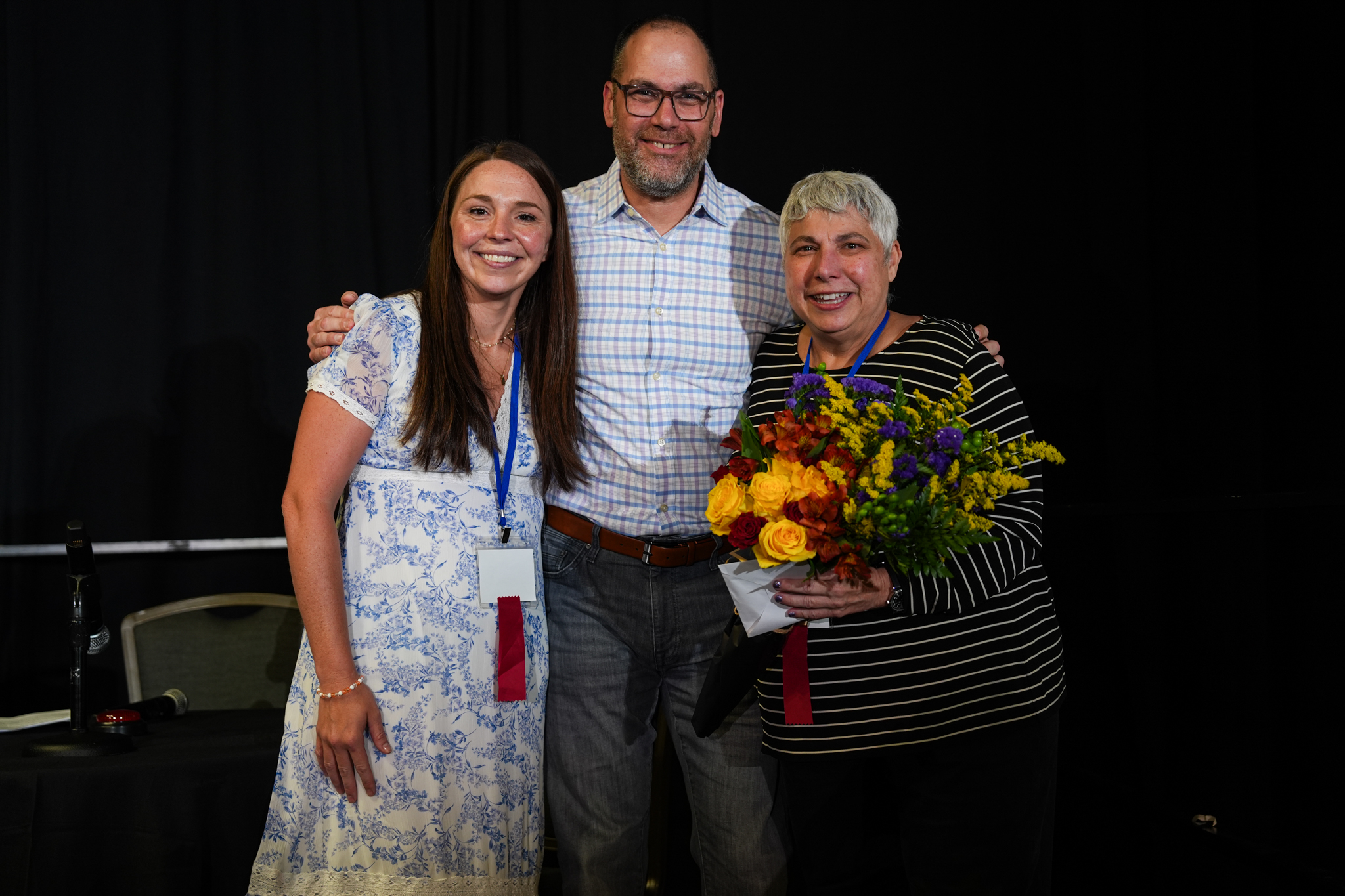 Three people smile together at a Vasculitis Foundation event. The woman on the right holds a colorful bouquet of flowers while the man in the center has his arms around both women.