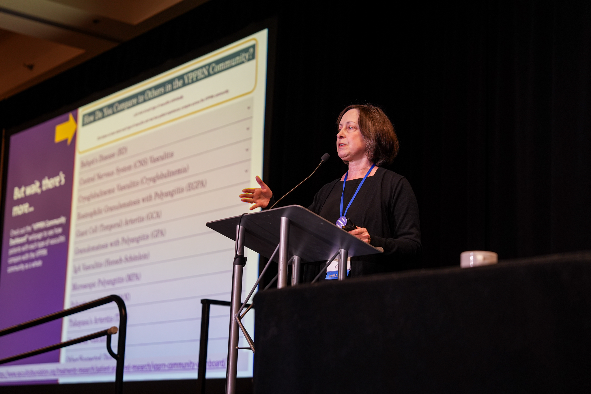 Dr. Jennifer Gordon speaks at a podium during the International Vasculitis Symposium, presenting information about the Vasculitis Patient-Powered Research Network (VPPRN) on a large screen behind her.