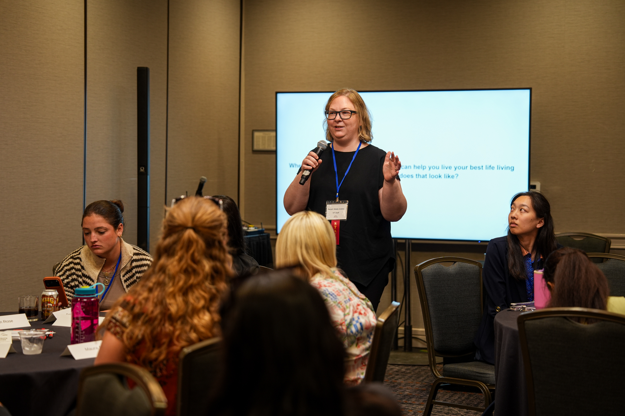A Vasculitis Foundation staff member speaks into a microphone during a small-group session at the International Vasculitis Symposium as attendees sit at tables around her.