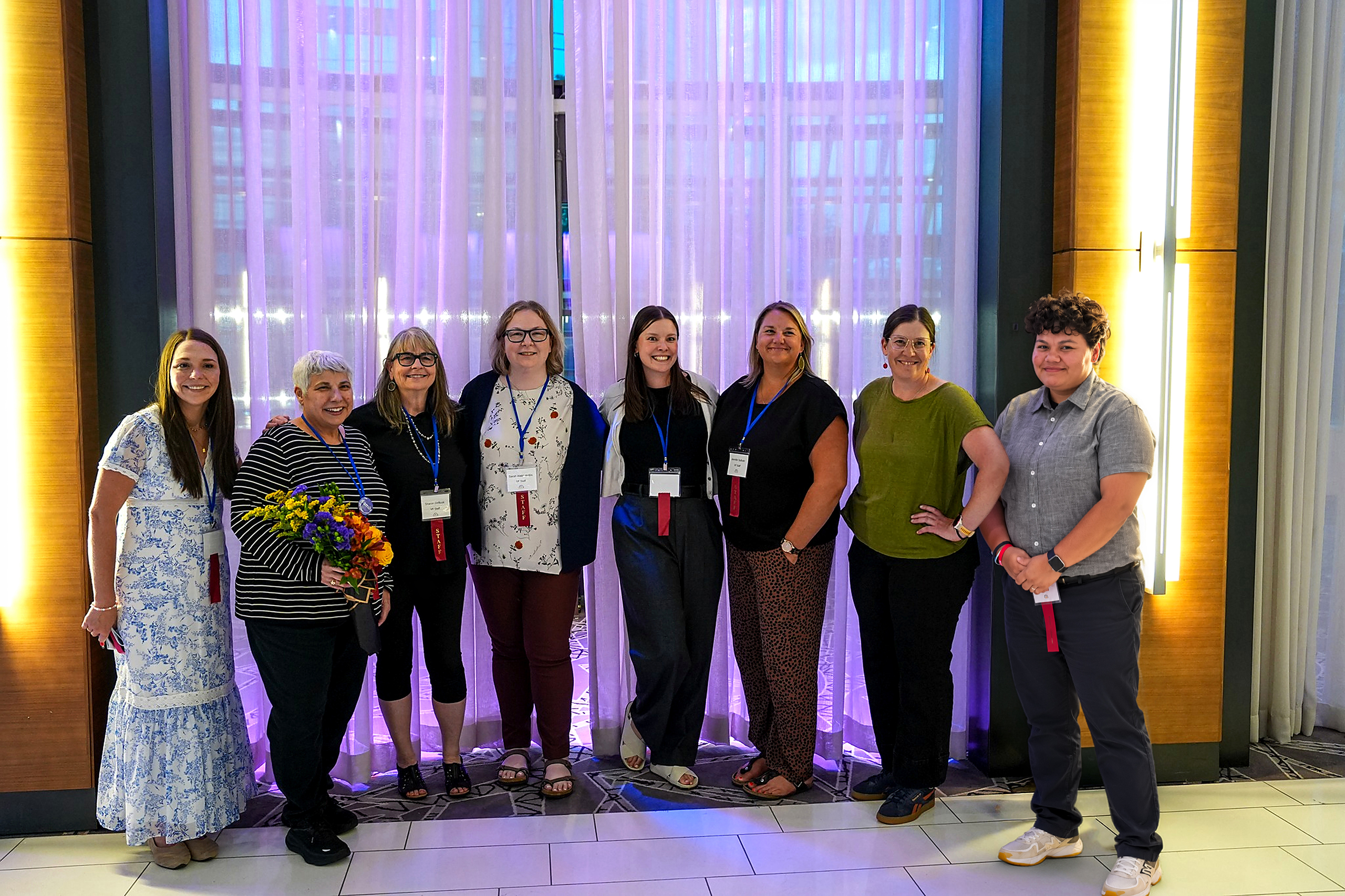 Eight Vasculitis Foundation staff members stand together and smile for a group photo at an event, with purple-lit curtains in the background. One person holds a colorful bouquet of flowers.