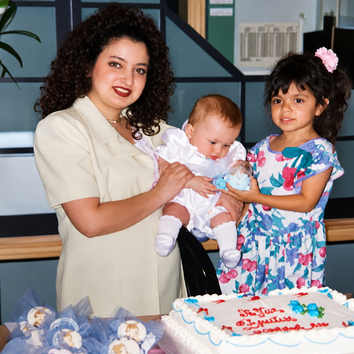 Archival family photograph of a woman holding a baby in a white outfit beside a young girl in a colorful floral dress, with a decorated cake in the foreground.