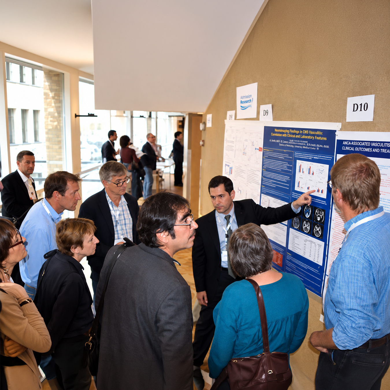 A presenter discusses vasculitis research findings while pointing to a scientific poster as a group of conference attendees gathers around to listen during a medical research poster session.