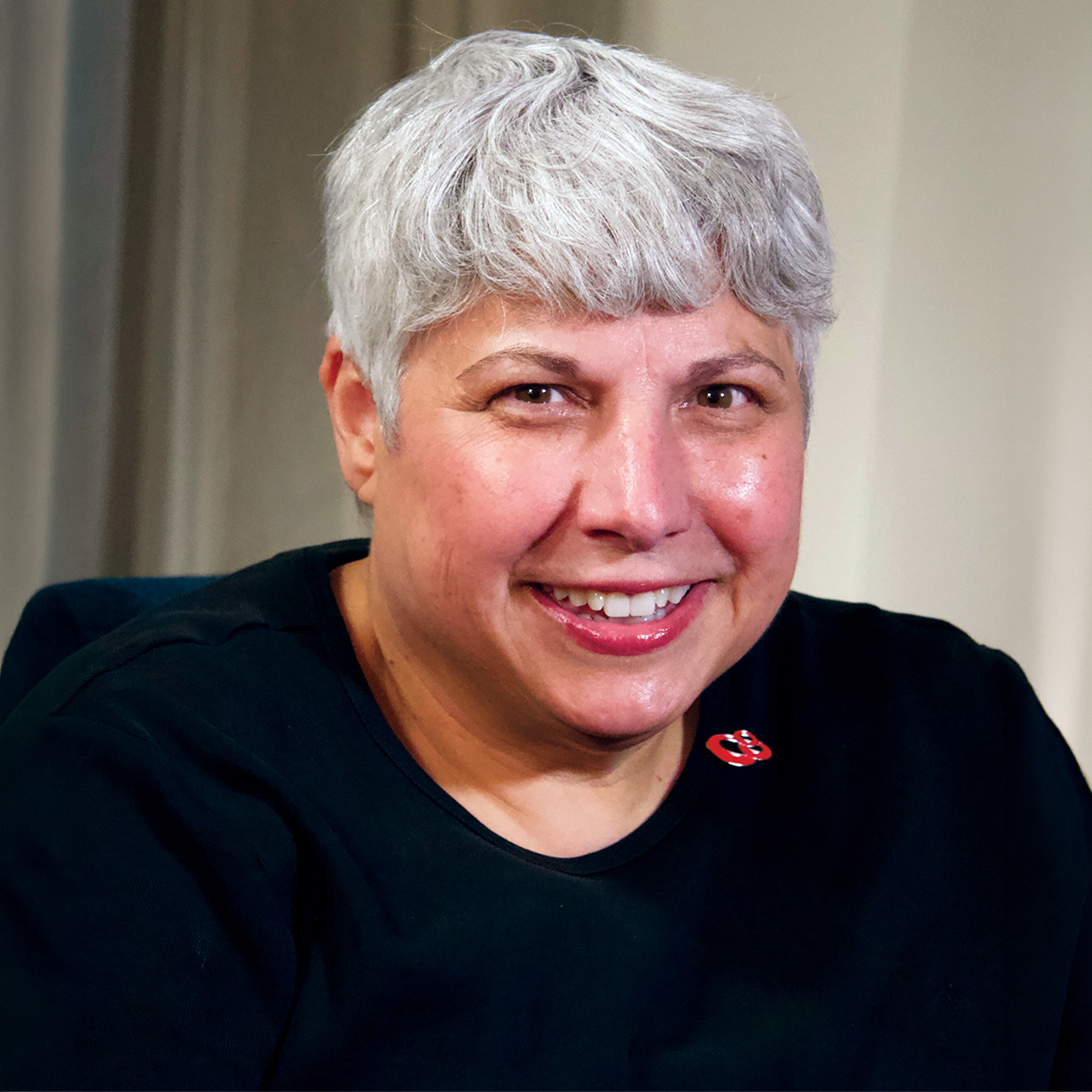 Portrait of Joyce Kullman smiling at the camera, wearing a black top with a small red pin and sporting short silver hair.