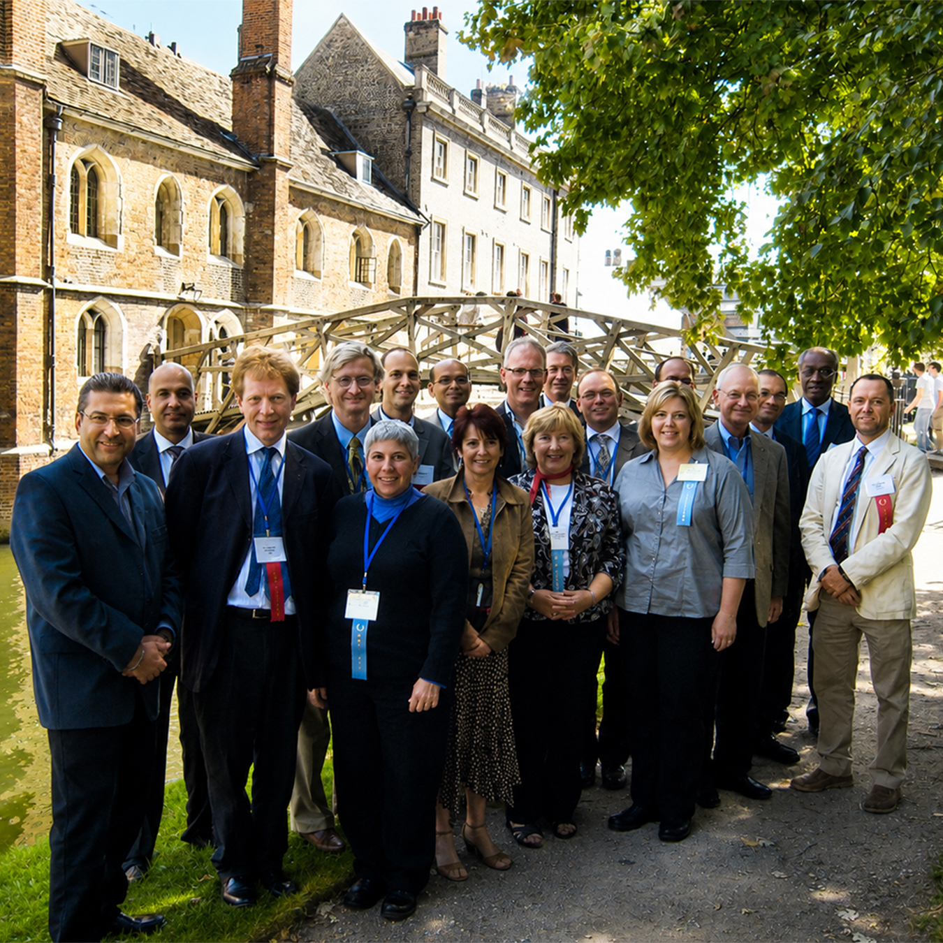 Group of symposium attendees and medical professionals wearing conference badges pose outdoors beside a historic stone building and wooden bridge in Cambridge, England.
