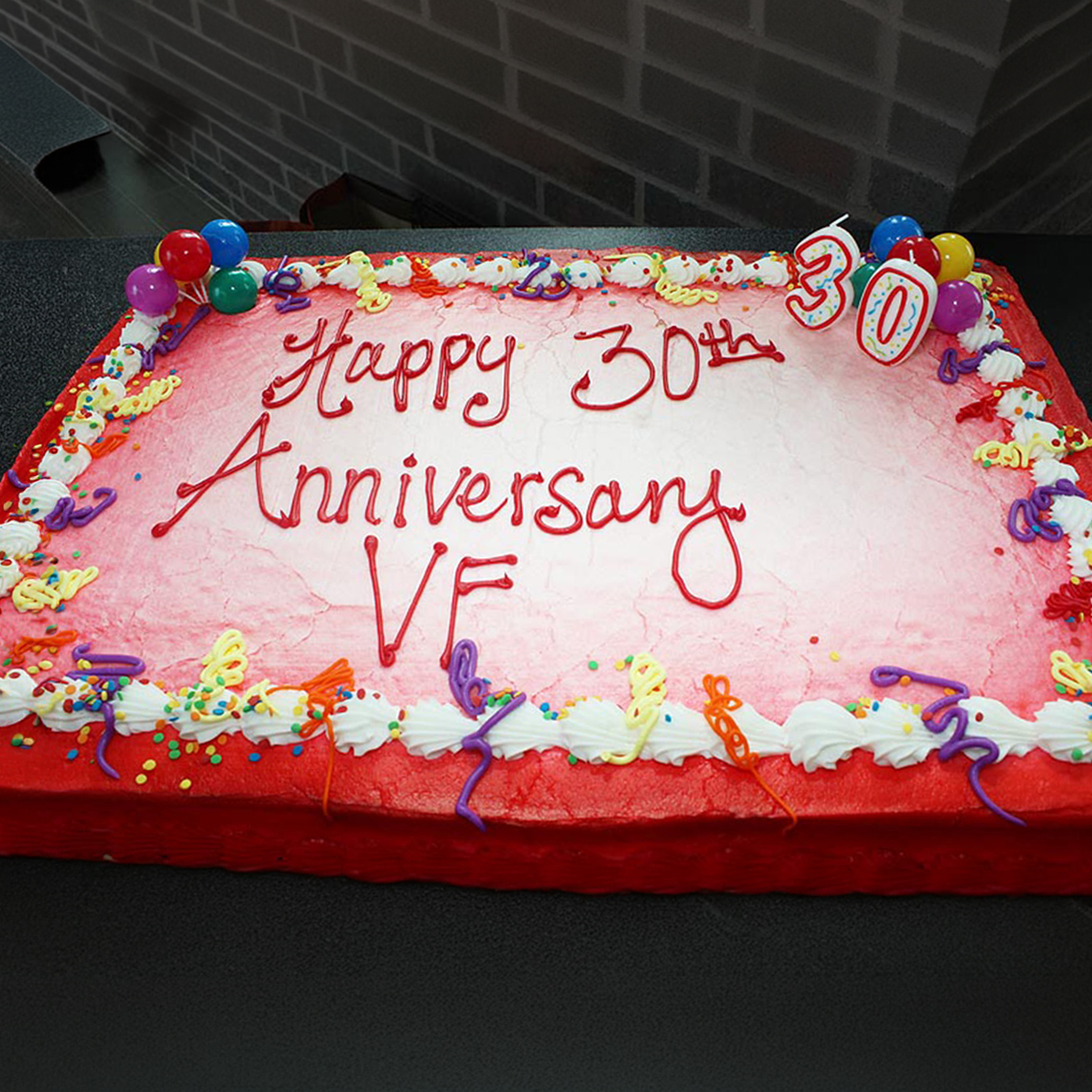 Large rectangular celebration cake decorated with pink and red frosting, colorful balloons, and the words “Happy 30th Anniversary VF.”