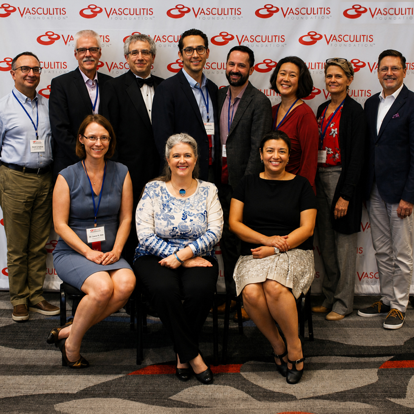 Eleven symposium presenters and medical professionals pose for a group photograph in front of a Vasculitis Foundation backdrop, with three seated in front.