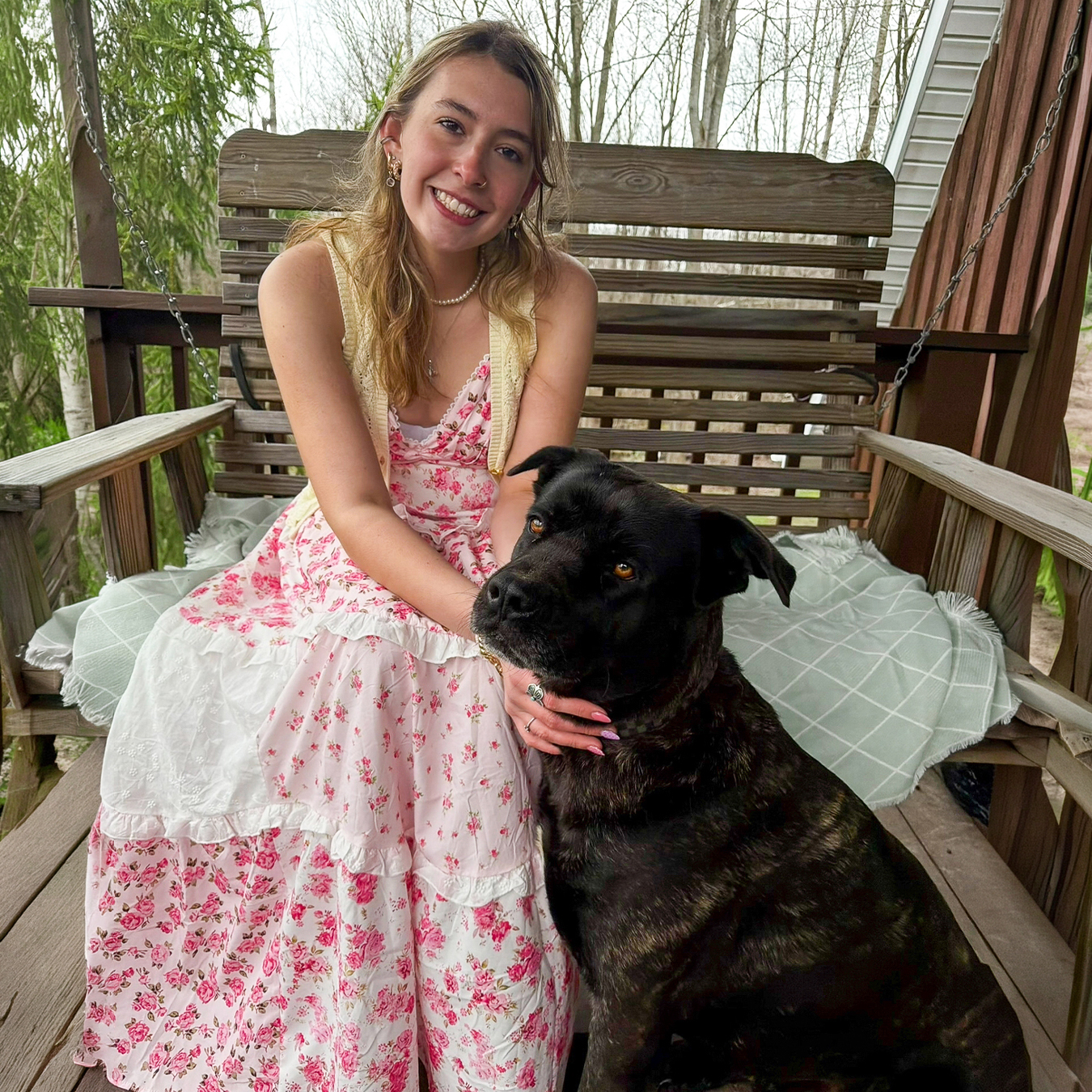 Chloe Cusano smiles while sitting on a wooden porch swing beside a large black dog, wearing a pink floral dress and a light yellow vest.