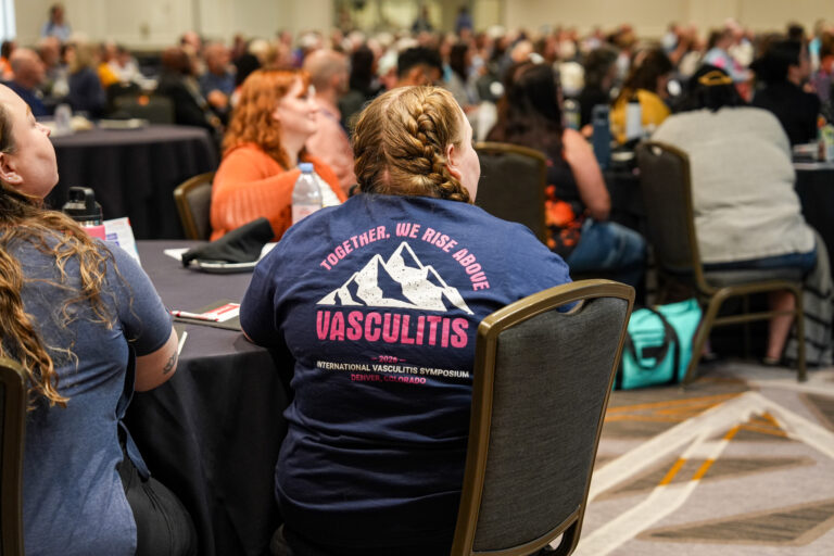 Attendees gather at the 2026 International Vasculitis Symposium in Denver, Colorado. In the foreground, a participant wears a navy shirt featuring a mountain design and the words “Together, We Rise Above Vasculitis.”