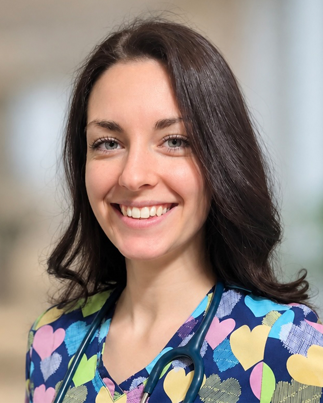 Professional headshot of a smiling woman with shoulder-length dark brown hair wearing colorful, heart-patterned medical scrubs and a stethoscope around her neck, against a softly blurred background.