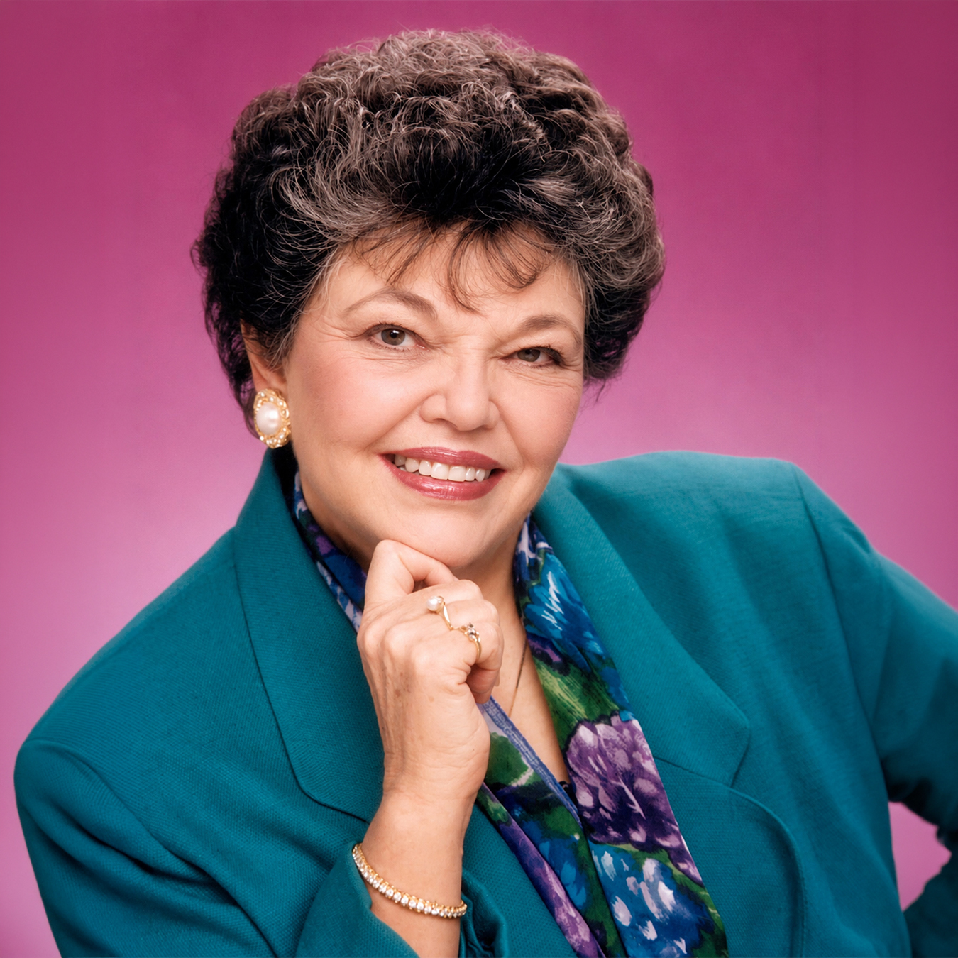 Studio portrait of Marilyn Samson smiling with her chin resting on her hand, wearing a teal blazer, colorful floral scarf, and pearl jewelry against a pink background.