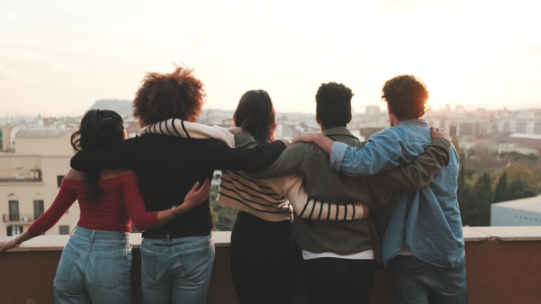 Five people stand with their arms around one another, overlooking a city skyline at sunset.