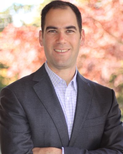 Professional headshot of a man wearing a dark blazer and light purple checkered shirt, smiling with a blurred outdoor background of autumn foliage.