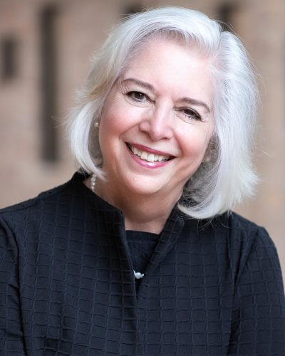 Professional headshot of a smiling woman with shoulder-length white hair wearing a black textured top, photographed against a softly blurred indoor background.