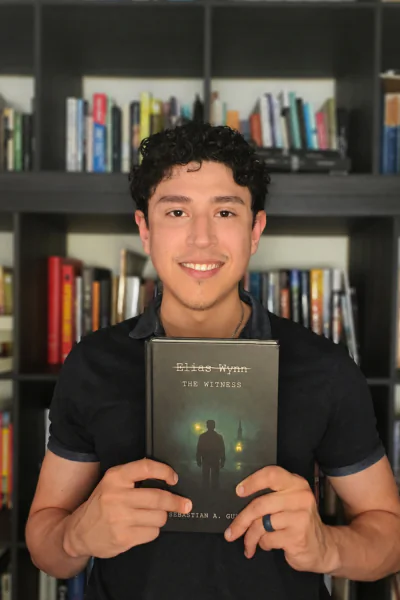 Sebastian pictured in front of book case holding "Elias Wynn THE WITNESS," a book he wrote.