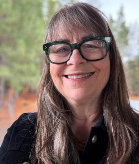 Smiling woman with long brown hair and glasses, standing outdoors in front of a blurred background