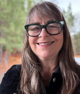 Smiling woman with long brown hair and glasses, standing outdoors in front of a blurred background