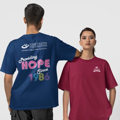 Shirt-Mockups Two people wearing Vasculitis Foundation T-shirts. The person on the left faces away, showing the back of a navy shirt with the Vasculitis Foundation logo and the words “Providing HOPE Since 1986” in bold retro-style lettering. The person on the right faces forward, wearing a maroon shirt with the Vasculitis Foundation logo on the chest.