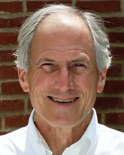 Smiling older man with gray hair wearing a white shirt, standing in front of a brick wall.
