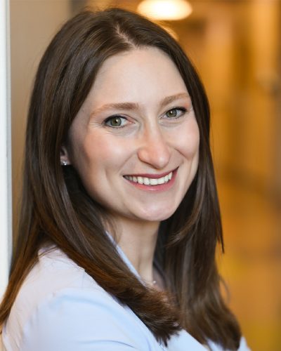Smiling woman with long brown hair in a light blouse, photographed indoors with a softly blurred background.