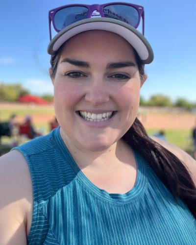 Smiling woman wearing a baseball cap and sunglasses on her head, outdoors at a field on a sunny day.
